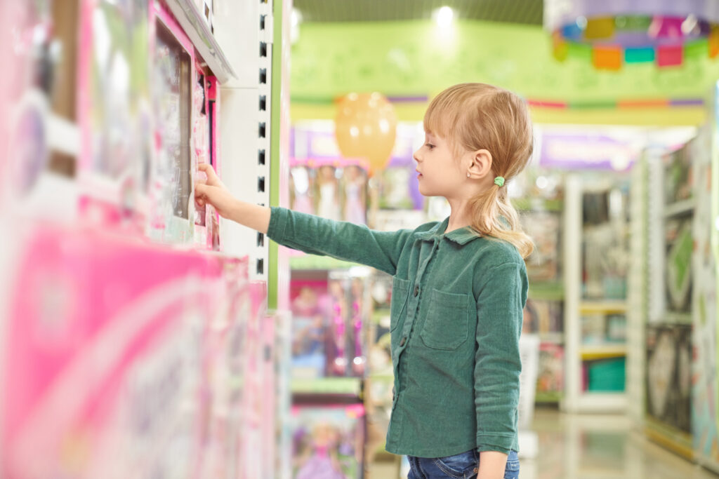 Pretty girl standing in store with children toys, looking at box with toy and taking from shelf. Big choice of children toys on shelves of modern shopping centre.